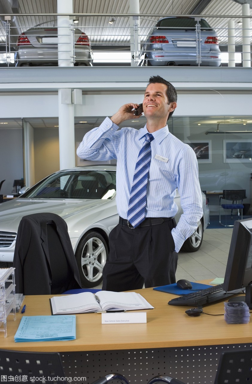 汽車(chē)銷(xiāo)售員站在柜臺(tái)旁的大型汽車(chē)陳列室里,用手機(jī)微笑著